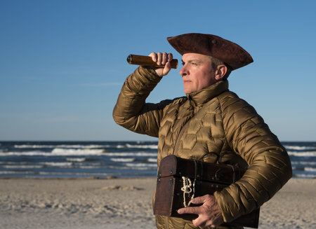 Man in pirate hat holds an ancient chest and looks intently into the distance through a spyglass....
