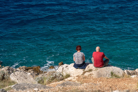 A man and a woman sit on a rocky seashore and enjoy the beautiful view of the blue, transparent sea.