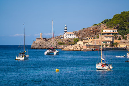 Picturesque view of a small lighthouse on pier in Port de Soller. Moored yachts, ships in harbor....