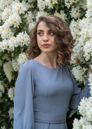 A beautiful girl with big eyes in a gray dress stands near white flowers. Grace, elegance, beauty,...