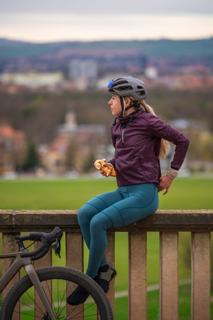 Cyclist sits on stone wall, eating pretzel and admiring panorama of Dresden in distance. Her gravel...