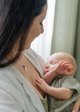Newborn baby sleeping soundly in mom loving arms. Woman standing by window, holding baby, stroking...