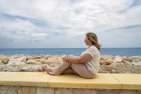 Side view of middle-aged woman sitting on pier of sea, reflecting and enjoying freedom and sensations of outdoors. Traveling and wanderlust way of life of people.の写真素材