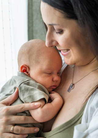 Woman tenderly holds her newborn son in arms. Mother looks at baby with warmth. Baby comfortably...