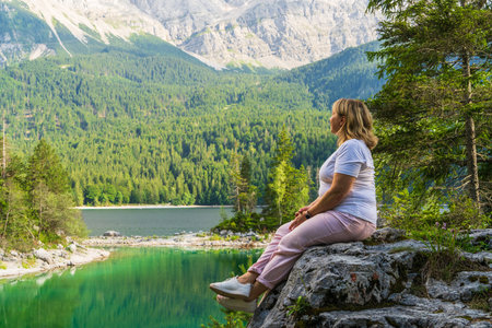 Female hiker rests on rock enjoying breathtaking views of Eibsee lake and surrounding mountains near...
