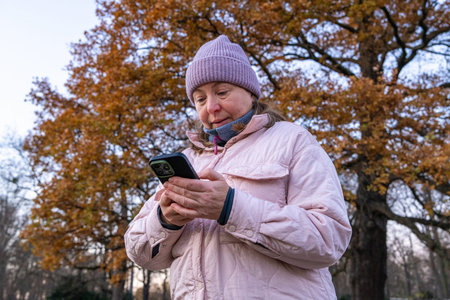 Middle aged woman walking in an autumn park, holding smartphone and dialing phone number to make...