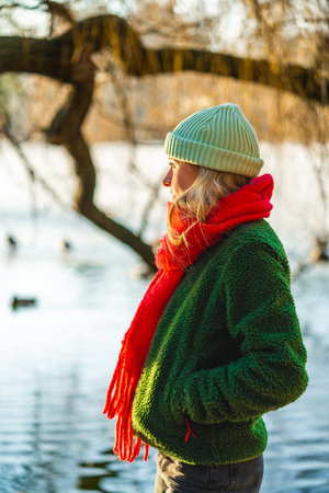 Modern young woman in green jacket and red scarf walking outdoors in park, hands in pockets, looking aside. Casual style, leisure, healthy lifestyle.の写真素材