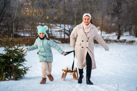 Smiling mother and little daughter running uphill with sled on snowy hill. Winter family leisure, happy childhood, parenting and active lifestyle.の写真素材