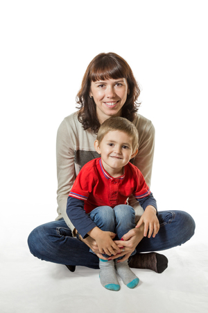 Smiling mother holding her son in her arms while sitting with him on the floor isolated on white backgroundの写真素材