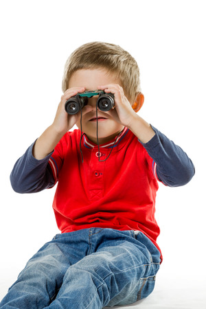 Little boy in blue jeans and a red sweater sitting on the floor and looking at the kids binoculars isolated on white backgroundの写真素材