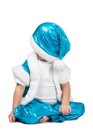 Kid in blue suit of Santa sitting on the floor and put his hat over his eyes isolated on white backgroundの写真素材