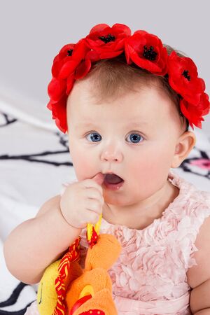 Baby girl with a wreath of red poppies on the head playing toy and takes the thumb in his mouthの写真素材
