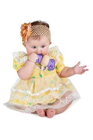 Baby girl in a dress and sling with a bow on head sitting and munches wooden toy isolated on white backgroundの写真素材