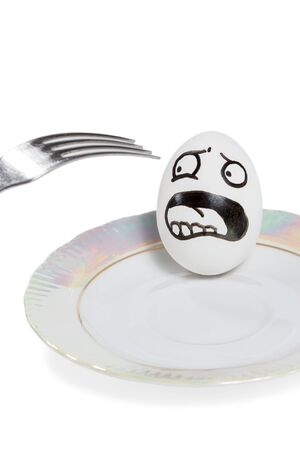 Egg with smiley lies in the plate and yells of fright because of approaching a fork isolated on a white backgroundの写真素材
