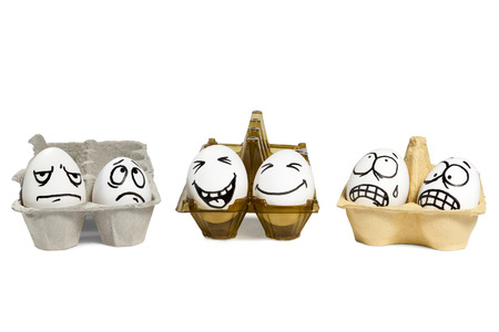 Two eggs with funny faces in a plastic tray, and next to them four eggs with angry faces in paper trays isolated on white backgroundの写真素材