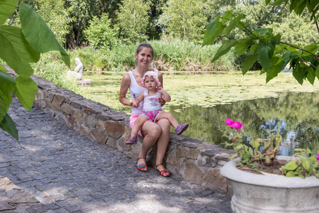 Portrait of a mother with a young daughter in her arms sitting on a curb stone at a pondの写真素材