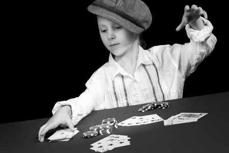 Child in a shirt and a cap pulled his hand to a deck of cards at a poker table with chips and cards, isolated on a black background, black and white photographyの写真素材