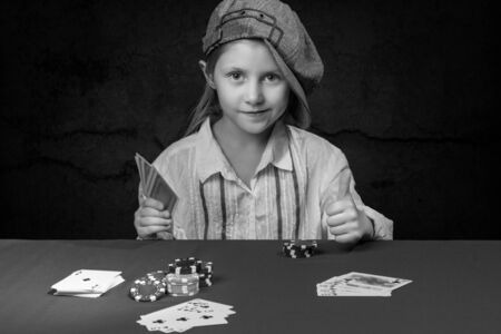 Smiling young girl in cap sitting at a poker table with cards in hand and shows lifted upwards thumb, black and white photoの写真素材