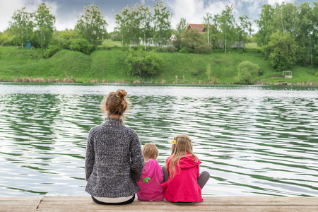 Mother with two daughters on a wooden board on the shore of lake rear viewの写真素材