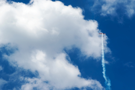 Small sports plane flies straight up into the blue sky with clouds and releasing smokeの写真素材