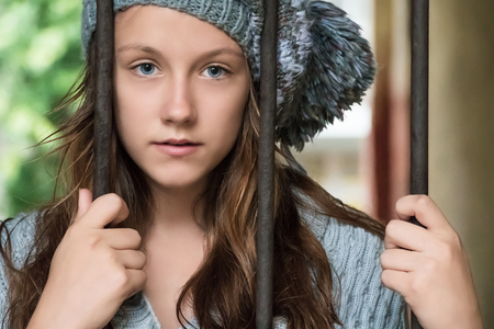 Close-up portrait of a beautiful young woman in a knitted cap and a sweater behind iron barsの写真素材