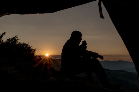 Silhouettes of tourists with a cup of tea in hand on background of mountains. The view from the open tentの写真素材