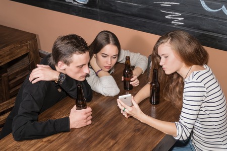 Three friends, two women and one man sitting in a cafe and look at phone screenの写真素材