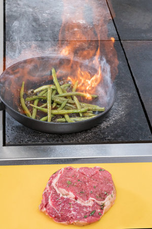Green asparagus cooked on a burning pan. Raw pork steak lies next on queue