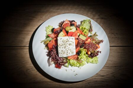 Beautifully decorated Greek salad with whole piece of feta cheese on top, served on a white plate on a wooden table in the darkの写真素材