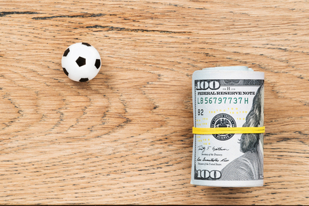 Little soccer ball with a roll of dollars lays on an old wooden tableの写真素材