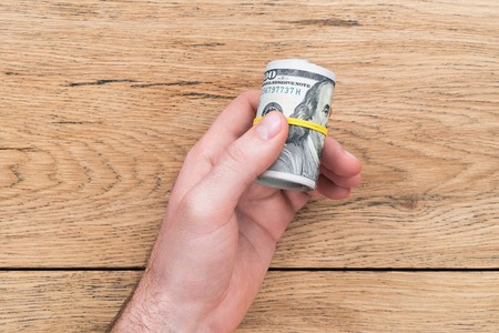 Male hand offering money - a roll of American dollars tied with yellow rubber band on the background of an old wooden tableの写真素材