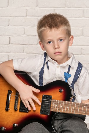Little beautiful boy playing on electric guitar sitting on a chair near brick wallの写真素材