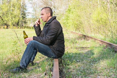 Lonely man with a cigarette and a bottle of beer sitting on rusted railsの写真素材