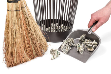 Conceptual photo - crumpled dollars on a garbage scoop next to a trash can and a broom isolated on a white backgroundの写真素材