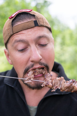 Bearded man eats a shish kebab tearing him from the skewer with his mouth, closeupの写真素材