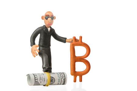 Bald plasticine businessman with glasses stepped on a roll of dollars and holds in his hand bitcoin isolated on white backgroundの写真素材