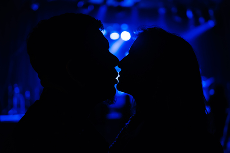 Silhouette of kissing man and woman on blurred background with blue lightsの写真素材