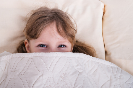 Little beautiful girl with blue eyes peeking out from under the blanketの写真素材