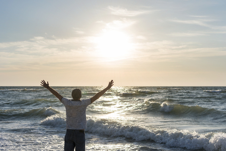 Portrait of a man with raised arms near the sea at dawn, standing with his back to the cameraの写真素材