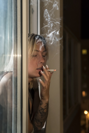 Woman is smoking a cigarette at night by the windowの写真素材