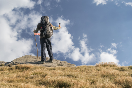 Tourist with a large backpack on his back stands on top of the mountain, holds an action camera in his hand and takes pictures of the skyの写真素材