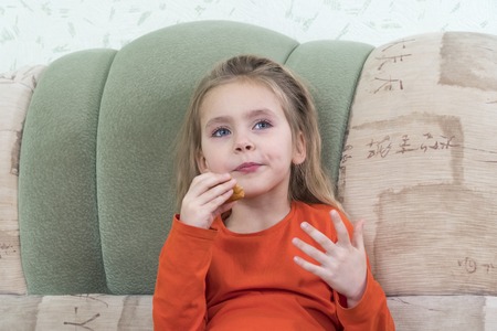 little girl happily eats bread sitting on the couchの写真素材