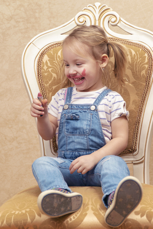 pretty child smeared with lipstick sits on a retro chair and looks at lipstickの写真素材