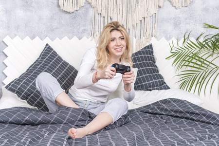 Young beautiful woman is enthusiastically playing a computer game with a joystick while sitting on a bedの写真素材