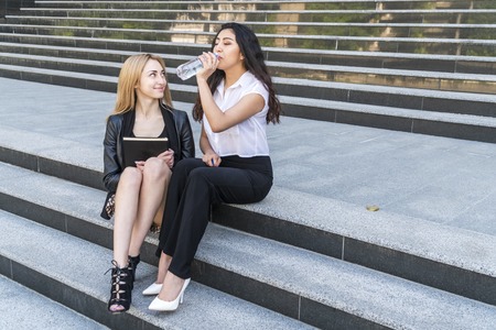 two female students drink water sitting on the steps of the university with a book in handの写真素材