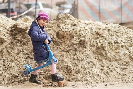 beautiful child carries a scooter on his shoulder, stepping over a stone near a slide of sandの写真素材