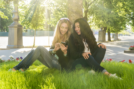 two beautiful women look into the phone with a smile while sitting on the grass by a treeの写真素材