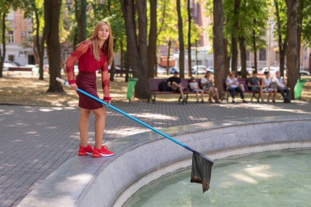 beautiful girl in red clothes collects trash from the surface of the water in a fountain in the park