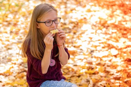 beautiful girl in glasses with an appetite eats an apple on a background of autumn foliageの写真素材