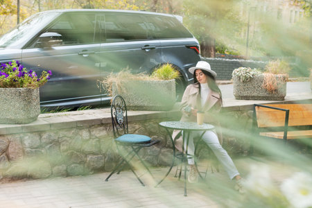 Woman with phone and a cup of coffee in a cafe on the street, shot through the foliage